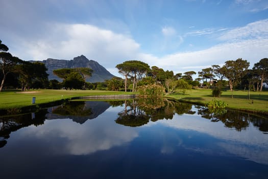 Grass, lake and trees on golf course with mountain, clouds and natural landscape with river in park. Nature, green and field with sustainable environment, sunshine and blue sky reflection in Canada.