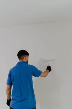 One young handsome dark-haired bearded Caucasian man in a blue t-shirt stands with his back and paints a wall with a roller of white paint, bottom side close-up view with selective focus and copy space.