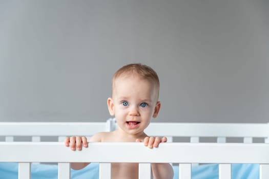 A modestly smiling baby leaned on the back of a wooden crib.