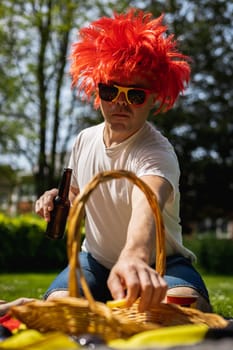 Portrait of a handsome young caucasian man in sunglasses, red wig holding a beer and reaching out with his hand for chips in a basket, celebrating belgium day at a picnic in a city park on a sunny summer day, close-up side view.