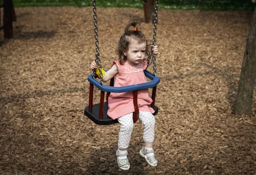 Portrait of one beautiful Caucasian baby girl in a pink dress with a ponytail on the top of her head with a sad and dissatisfied emotion on her face rides on a swing on a summer day in the park on the playground, side view close-up.