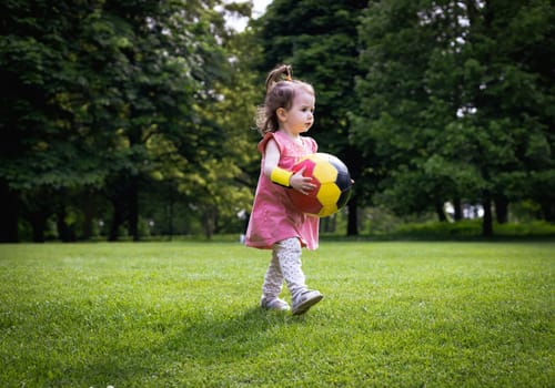 A little Caucasian girl in a dress is holding a soccer ball while running on the green lawn of a city park on a summer day, close-up view from the side.