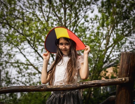 Portrait of one beautiful Caucasian brunette girl with glasses and a Belgian flag hat, looking at the camera with a smile and standing on a wooden bridge on a playground in a city park, close-up view from below.