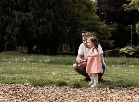 Portrait of one beautiful Caucasian little girl in a pink dress with a ponytail on the top of her head standing near her father squatting and carefully looking away in the park on a summer day, close-up side view with copy space on the left.