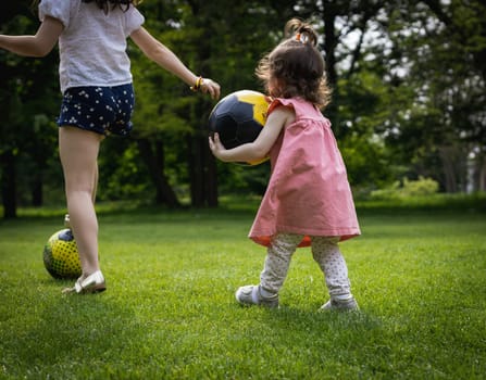 Two small Caucasian girls from the back playing with a soccer ball on a green lawn in a city park on a summer day, close-up view from the side.
