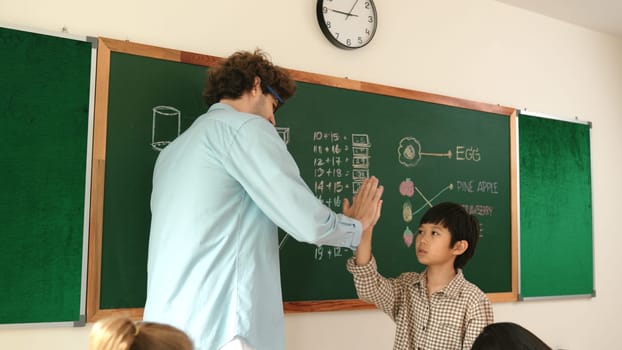 Attractive boy writing answer while teacher give high five to encourage child. Attractive student walking back to his seat while teacher applause and clap hand to schoolboy at Math lesson. Pedagogy.