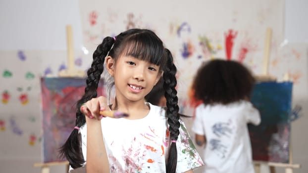 Creative cute girl pose at camera while diverse children painting behind. Young beautiful child looking at camera while standing at stained room and holding paintbrush. Creative activity. Erudition.