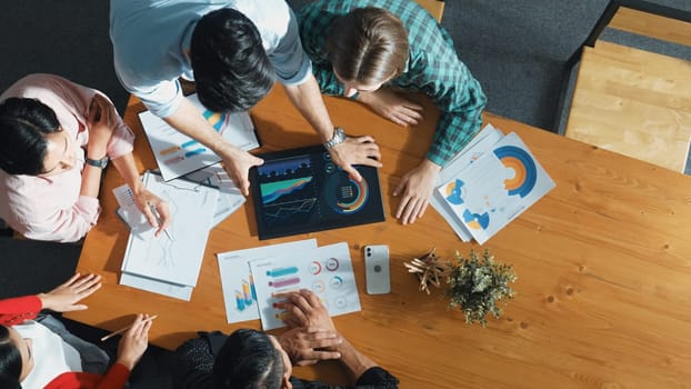 Top view of business man using tablet display increasing graph while diverse marketing people sharing idea or planing strategy at meeting table with stock market statistic scatter around. Convocation.