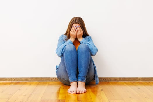 Upset young woman sitting on wooden floor with hands covering face. Emotional well-being concept with copy space for design and print.