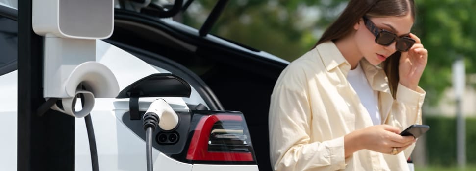 Young woman use smartphone to pay for electricity at public EV car charging station green city park. Modern environmental and sustainable urban lifestyle with EV vehicle. Panorama Expedient