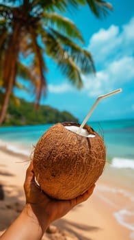Hand holding a coconut with a straw against a tropical beach backdrop