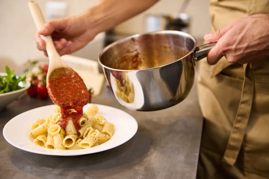 Hands of a man holding a stainless steel pan and pouring tomato sauce on pasta, cooking dinner at home kitchen