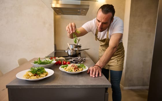 Attractive European male chef decorating spaghetti Bolognese with fresh greens, standing at kitchen island in the domestic kitchen. Man cooking dinner at home. Culinary. Italian food concept