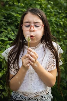 Portrait of one beautiful Caucasian brunette girl in glasses emotionally and strongly blowing on a dandelion, standing in the park on a summer day, close-up side view.