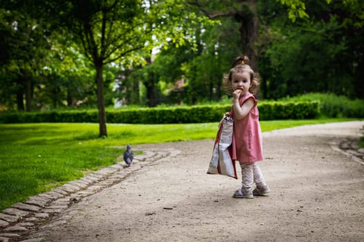 Portrait of one beautiful Caucasian girl in a pink dress with a ponytail on the top of her head standing in the park and eating chips from a large bag, close-up side view.