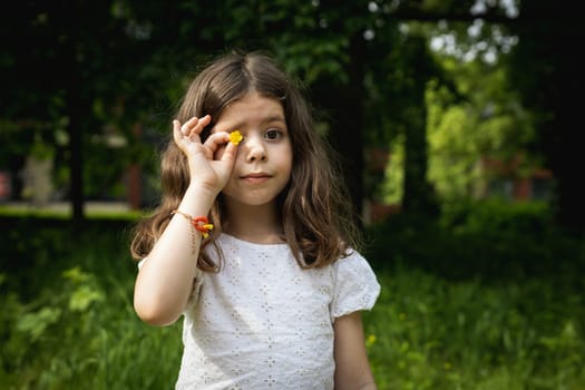 Portrait of a beautiful brunette Caucasian girl with loose hair holding a yellow flower over her right eye in the park on a summer day.