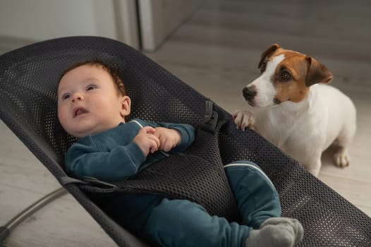 A dog sits next to a cute three-month-old boy dressed in a blue overalls in a baby lounger
