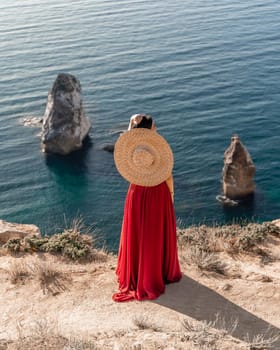 A woman in a red dress stands on a rocky cliff overlooking the ocean. She is wearing a straw hat and she is enjoying the view