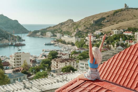 Woman sits on rooftop with outstretched arms, enjoys town view and sea mountains. Peaceful rooftop relaxation. Below her, there is a town with several boats visible in the water.