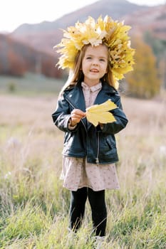 Little smiling girl with a yellow leaf in her hand and a wreath of autumn leaves stands in a field. High quality photo