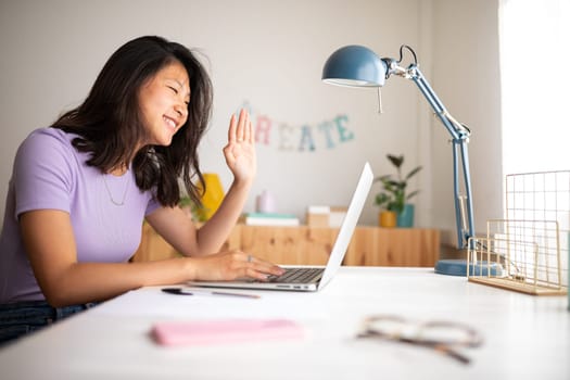 Chinese teen college student in video call taking online classes. Greeting classmates. Waving hand towards laptop. E-learning concept. Working from home. Video call concept.