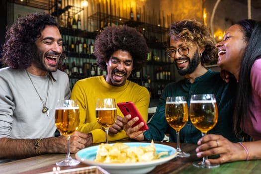 Group of multiracial friends laughing looking mobile phone together. Having fun in a bar drinking beer. Lifestyle and technology concept.