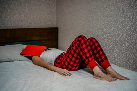 Young brunette woman works and studies from home on a laptop, while lying on her bed in red checkered pants. Balancing work and studies at home