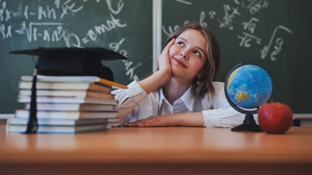 Adorable school girl posing at her desk against a background of blackboard, books, globe and graduation cap