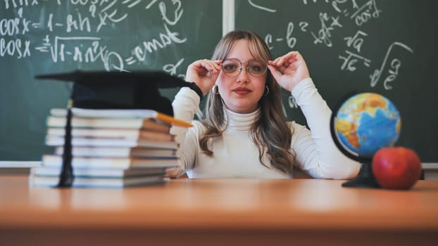 A schoolgirl wearing glasses poses against a background of books, an apple, a globe and a graduation cap