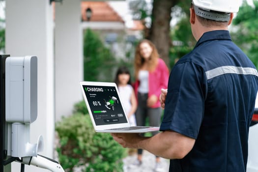 Qualified technician working on home EV charging station installation, making troubleshooting and configuration setup on charging system with laptop for EV at home with the family present. Synchronos