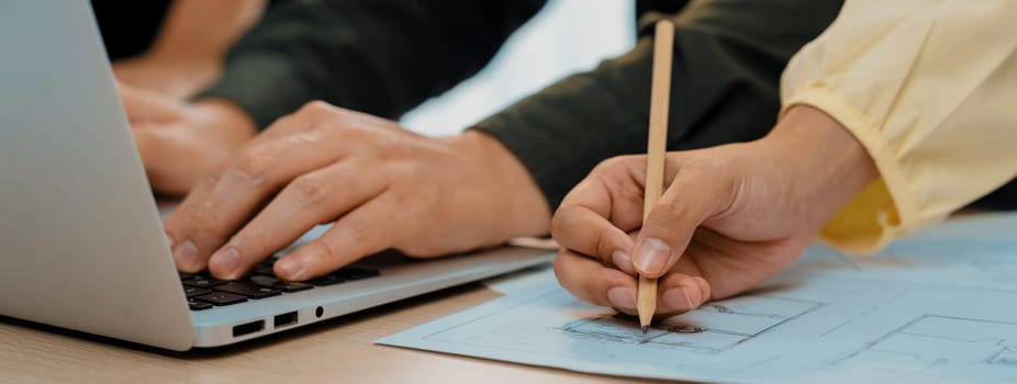 A portrait of skilled architect engineer hand writing a detail of blueprint during male engineer searching data from laptop at modern architectural office. Focus on hand. Closeup. Delineation.