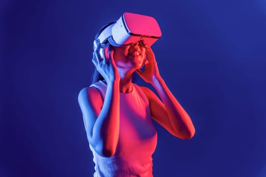 Smart female stand with surrounded by cyberpunk neon light wear VR headset connecting metaverse, futuristic cyberspace community technology. Elegant woman look generated virtual area. Hallucination.