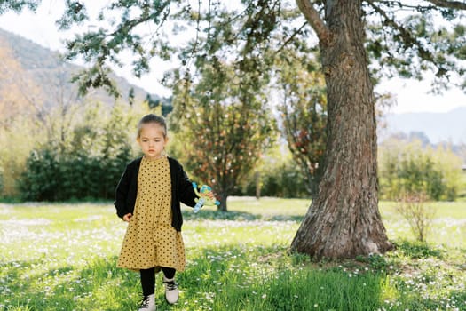 Little girl with a toy in her hand walks through a flowering meadow near a pine tree. High quality photo