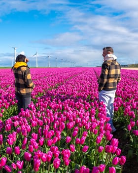 A man and woman standing tall amidst a sea of vibrant purple tulips in a stunning field in the Netherlands, with windmill turbines in the background symbolizing the beauty of Spring.