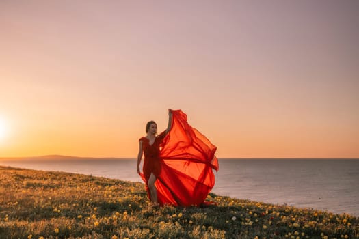 A woman in a red dress is standing on a grassy hill overlooking the ocean. The sky is a beautiful mix of orange and pink hues, creating a serene and romantic atmosphere