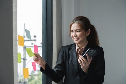 Young creative woman professional using sticky notes in glass wall to writing strategy business plan to development grow to success. Happy asian woman working at office..