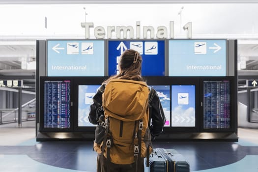 A woman with a backpack is looking at a large screen with a sign that says Terminal 1. She is looking at the screen with a sense of anticipation and excitement, possibly waiting for her flight