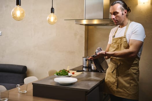 Handsome European man chef wiping wet hands with kitchen towel while cooking dinner at home kitchen. People and domestic life. Culinary concept