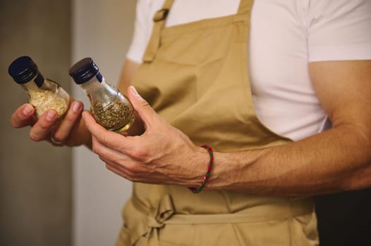 Close-up chef hands holding two glass bottles with condiments, dried basil leaves and ail for seasoning dish while cooking dinner