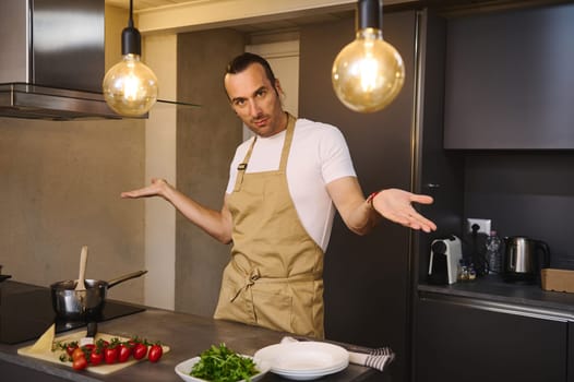Handsome Caucasian man in chef's apron cooking family dinner, smiling looking at camera, standing at kitchen counter with fresh ingredients in the modern minimalist home kitchen interior