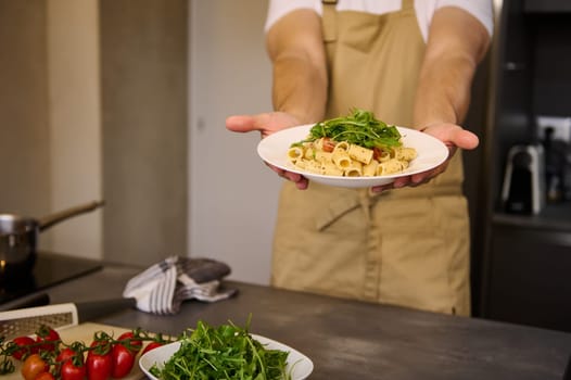 White plate with Italian pasta, garnished with tomato sauce and fresh arugula leaves in the hands of male chef holding out at camera the dish, standing at kitchen table with ingredients. Food concept.