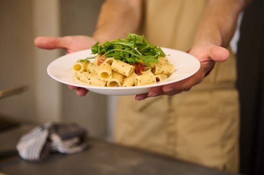 Selective focus on male chef hands holding a white plate with Italian pasta with tomato sauce, seasoned with basil and arugula leaves on chef's hands. Italian cuisine. Culinary. Food background