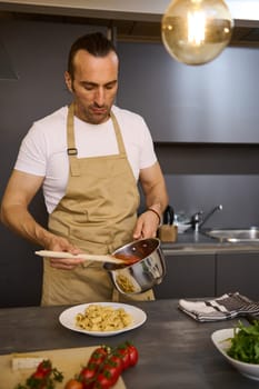 Vertical shot of a handsome European man in chef apron, pouring tomato sauce on Italian pasta, plating up the dish before serving. Man cooking dinner at home kitchen. Cuisine. Culinary. Epicure. Food