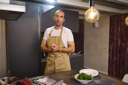 Authentic portrait of a handsome Caucasian male chef in beige kitchen apron, standing at kitchen counter with fresh ingredients, looking at camera, preparing dinner at home kitchen