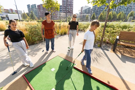 A woman is teaching her daughter to play golf. They are standing on the golf course, the girl is holding a golf club, the woman is standing behind her and directs her to strike. High quality photo