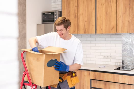 Smiling male worker of moving and delivery company holding cardboard box. Loader in overalls posing against background of colleague who packs cardboard boxes. Moving service concept