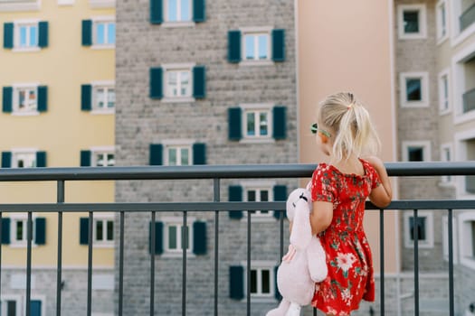 Little girl with a pink toy hare stands by the fence and looks at a colorful residential building. Back view. High quality photo