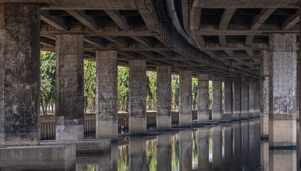 Bangkok, Thailand - Apr 20, 2024 - Perspective view of concrete pillars under Khlong Toei Expressway, with calm surface water reflection wonderful shadow, shape, lines and curve the underside support beams of the road along the Khlong phra khanong canal in Bangkok Thailand, Space for text, Selective focus.