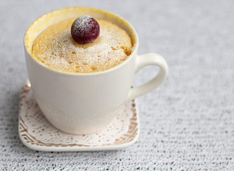 muffin in a mug decorated with cherries. a cup of white flour cake, sprinkled with powder, stands on a saucer, on a light background. Dessert in a mug. Microwave sweet pastries. High quality photo