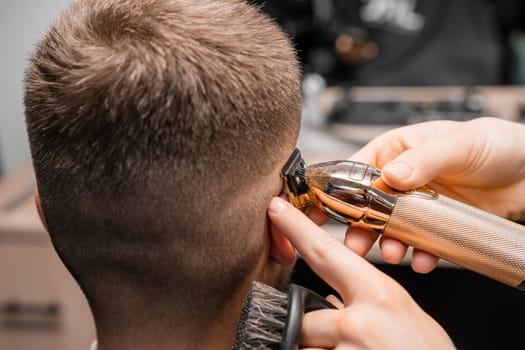 Barber shaves male client hair with trimmer in barbershop. Hairdresser shaving a mans temple